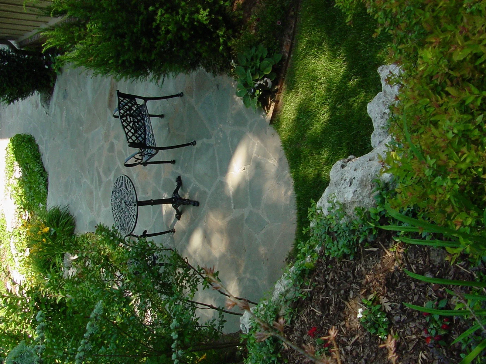 A backyard patio with a stone floor, a black metal bench, and a small black metal table, surrounded by green bushes and plants.