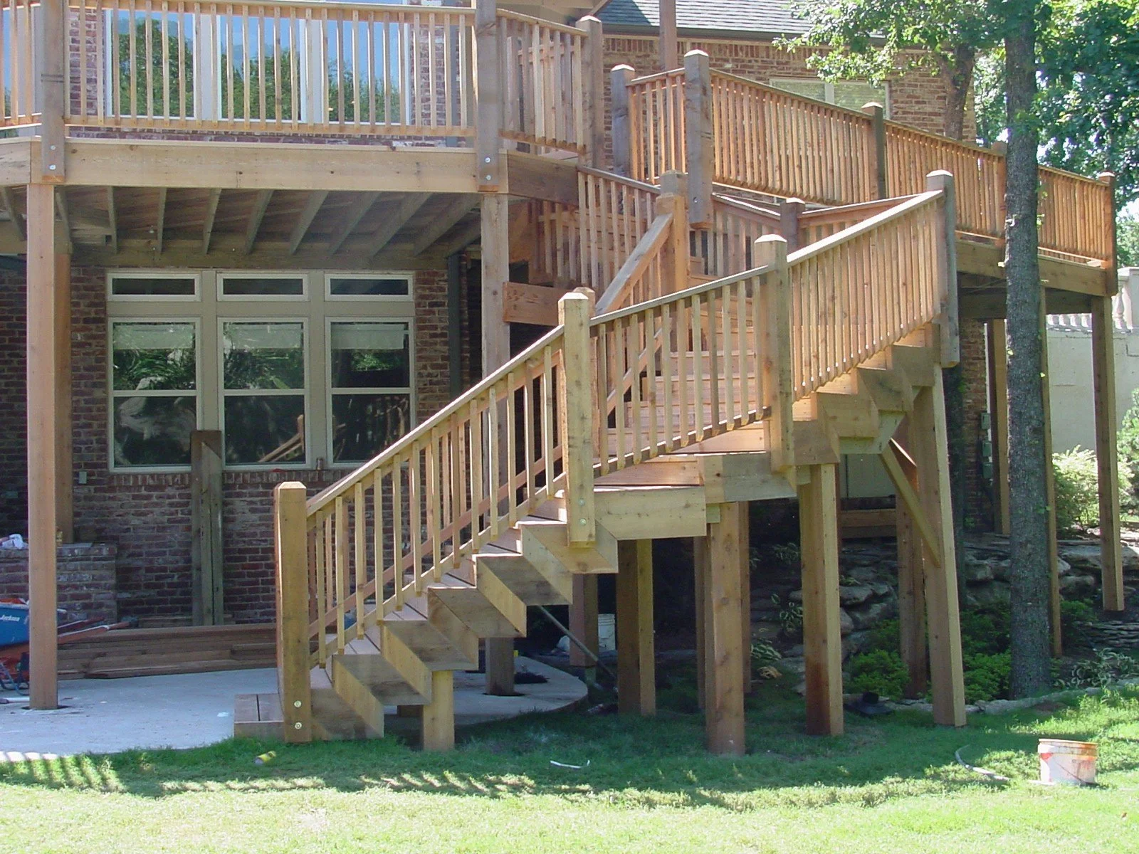 Newly constructed wooden deck with stairs attached to the back of a brick house, surrounded by trees and green lawn.