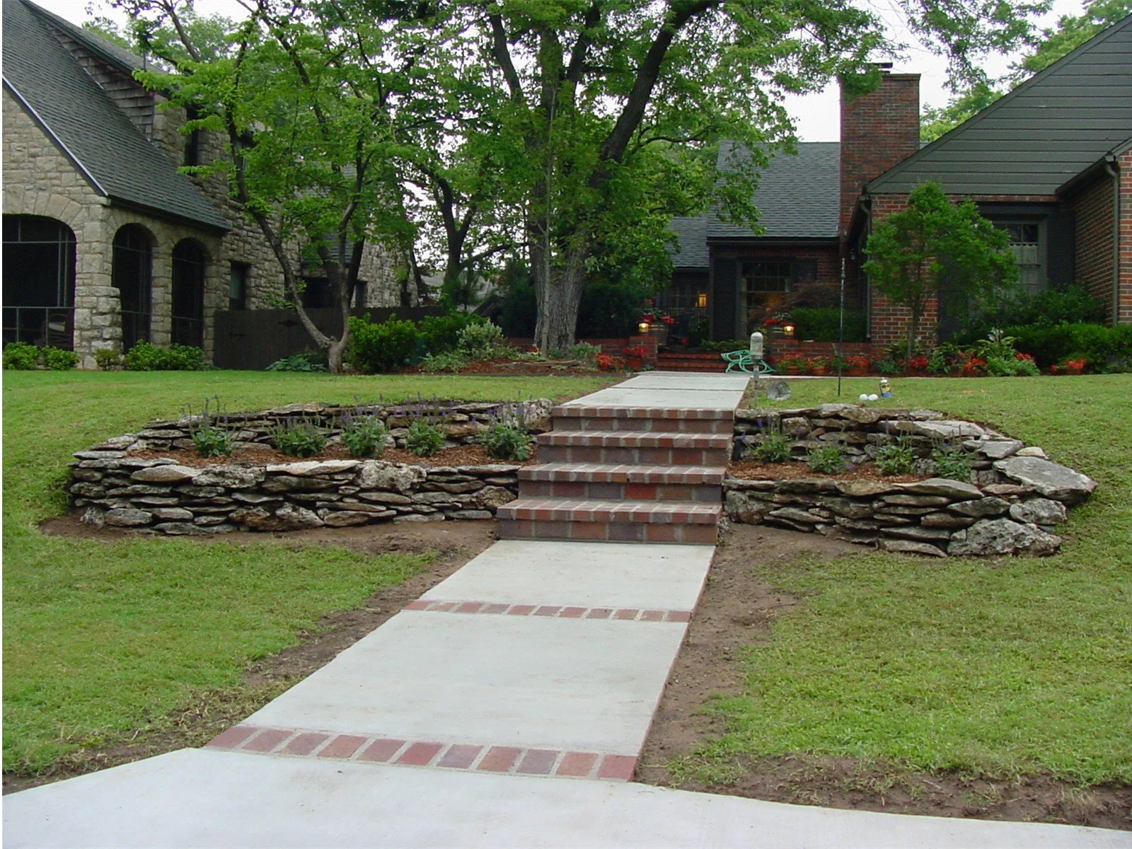A backyard with a brick pathway and stairs leading up to a patio, surrounded by a stone planter with plants, lush green grass, and large trees. There are houses in the background with stone and brick exteriors.