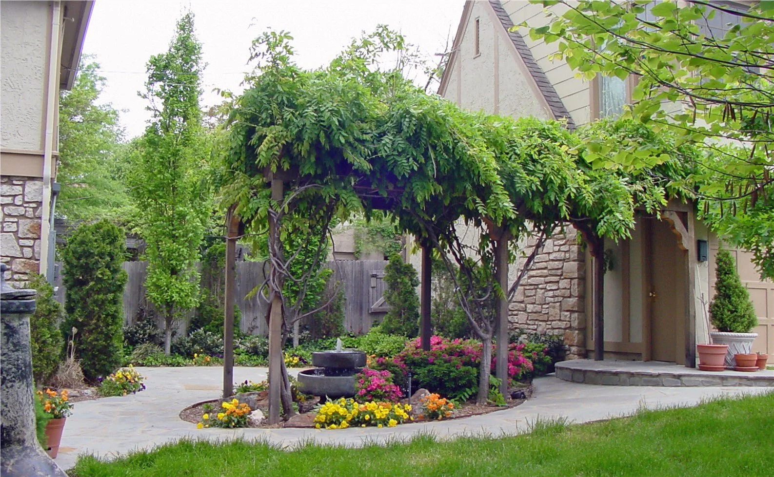 Residential front yard with a stone and wood exterior, lush greenery, a willow tree with a trellis, colorful flowers, potted plants, and a small curved patio with a stone fountain.