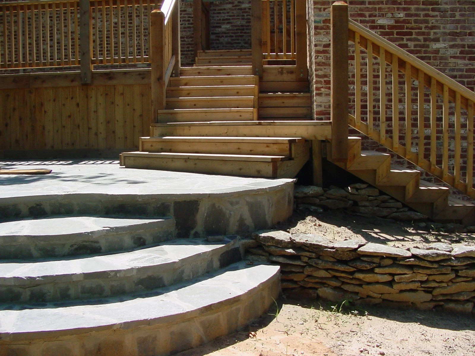 Wooden stairs leading to a deck, with a stone pathway and stone steps in the foreground, and a brick wall in the background.