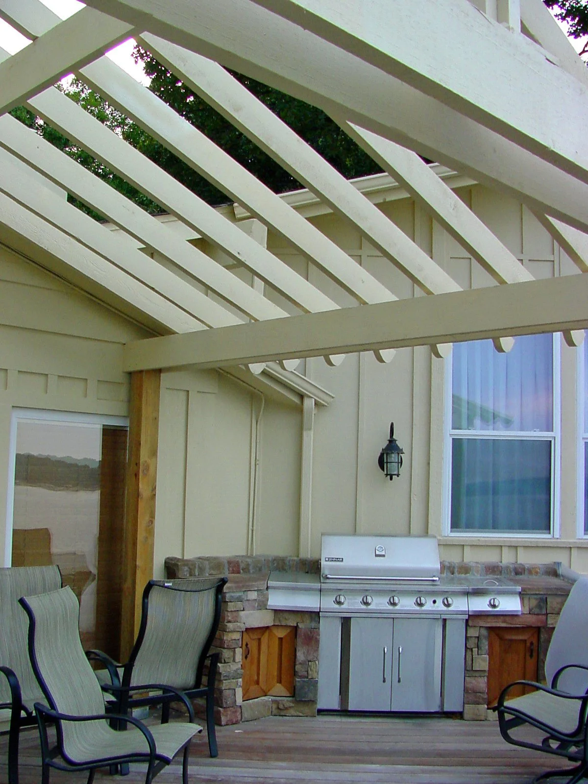 Outdoor patio area with a built-in barbecue grill, chairs, and a partially constructed pergola with white wooden beams.