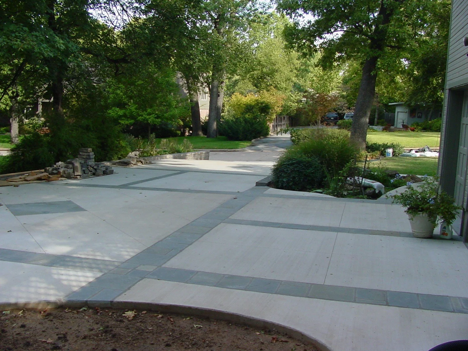 A newly paved concrete driveway with decorative gray stone borders, leading to a suburban yard with trees and grass, and a house on the right side.