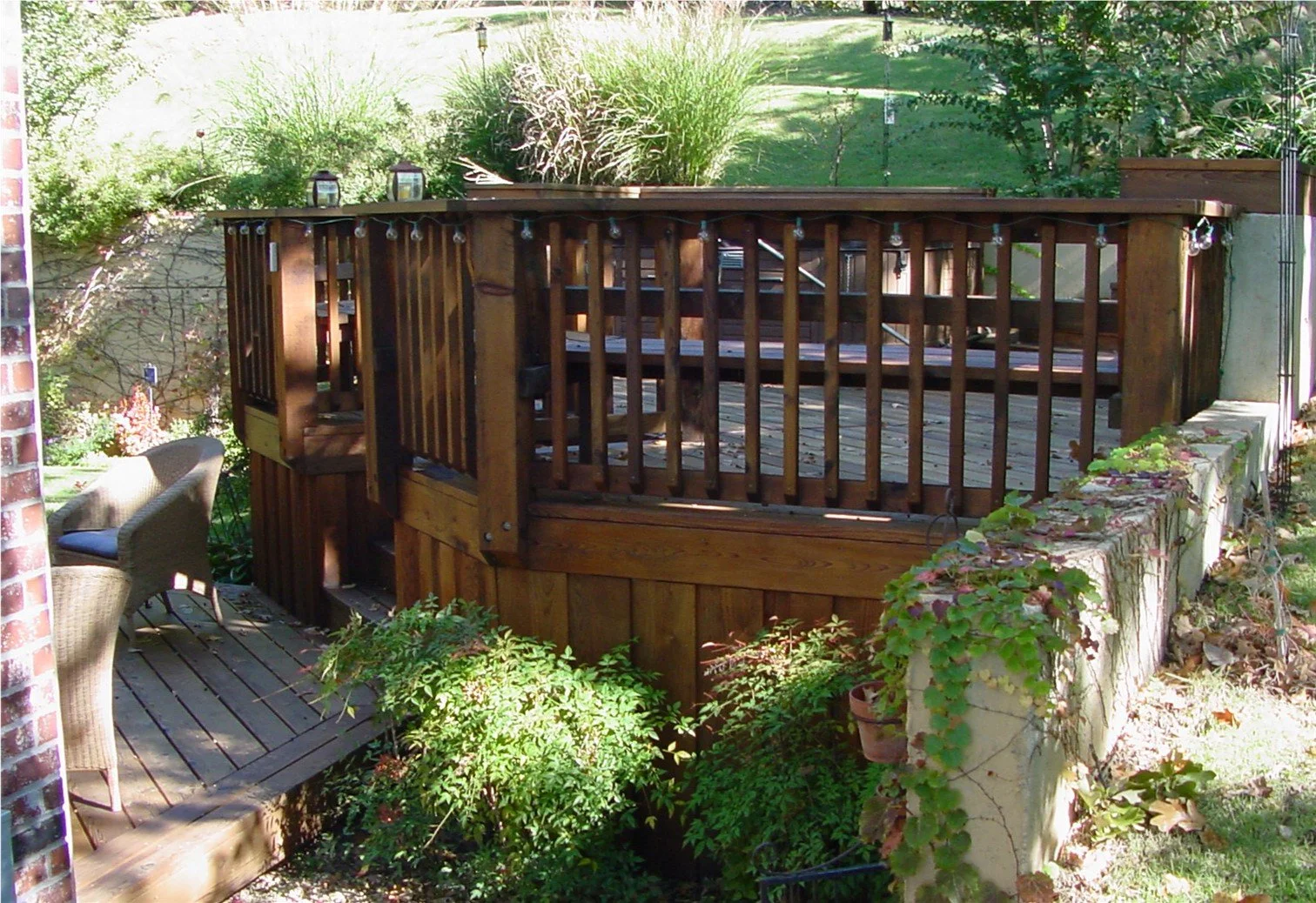 Wooden backyard deck with railing, outdoor seating chairs, and lush greenery and plants surrounding it.