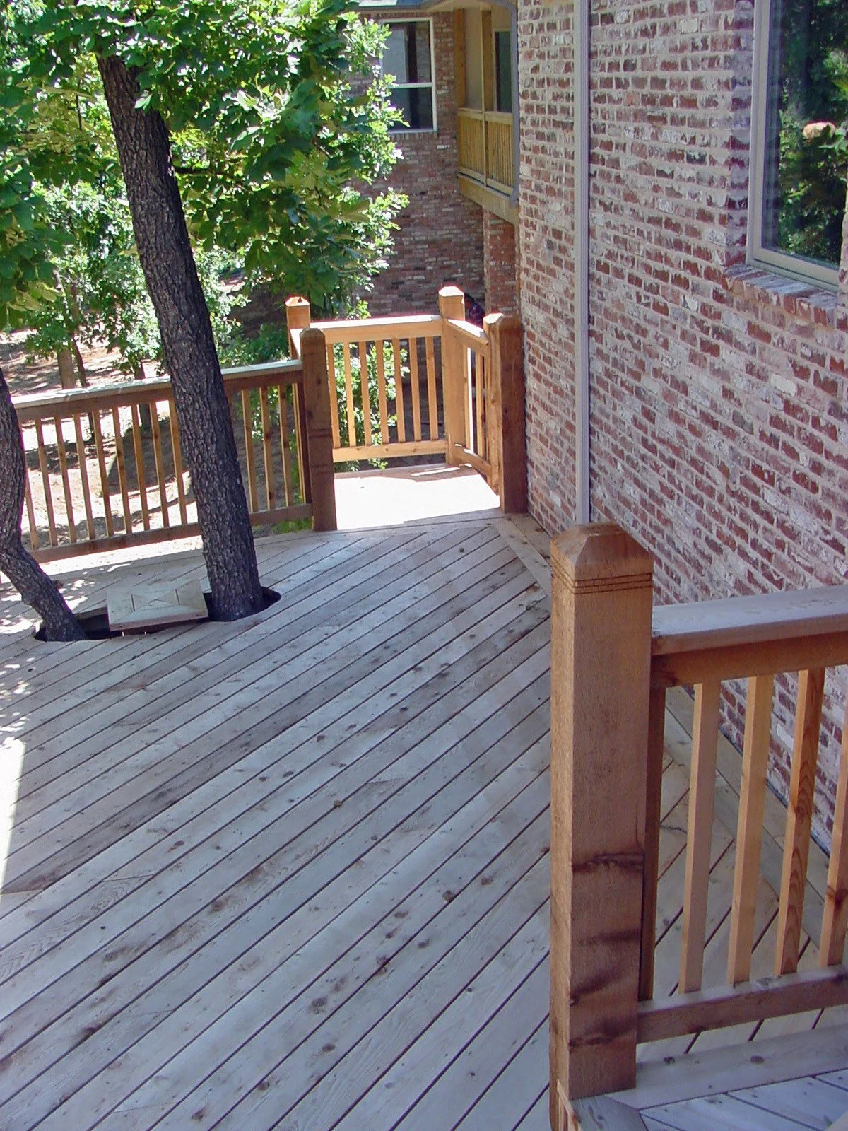 Wooden porch with a railing, brick exterior wall of a house, trees, and a small gate.