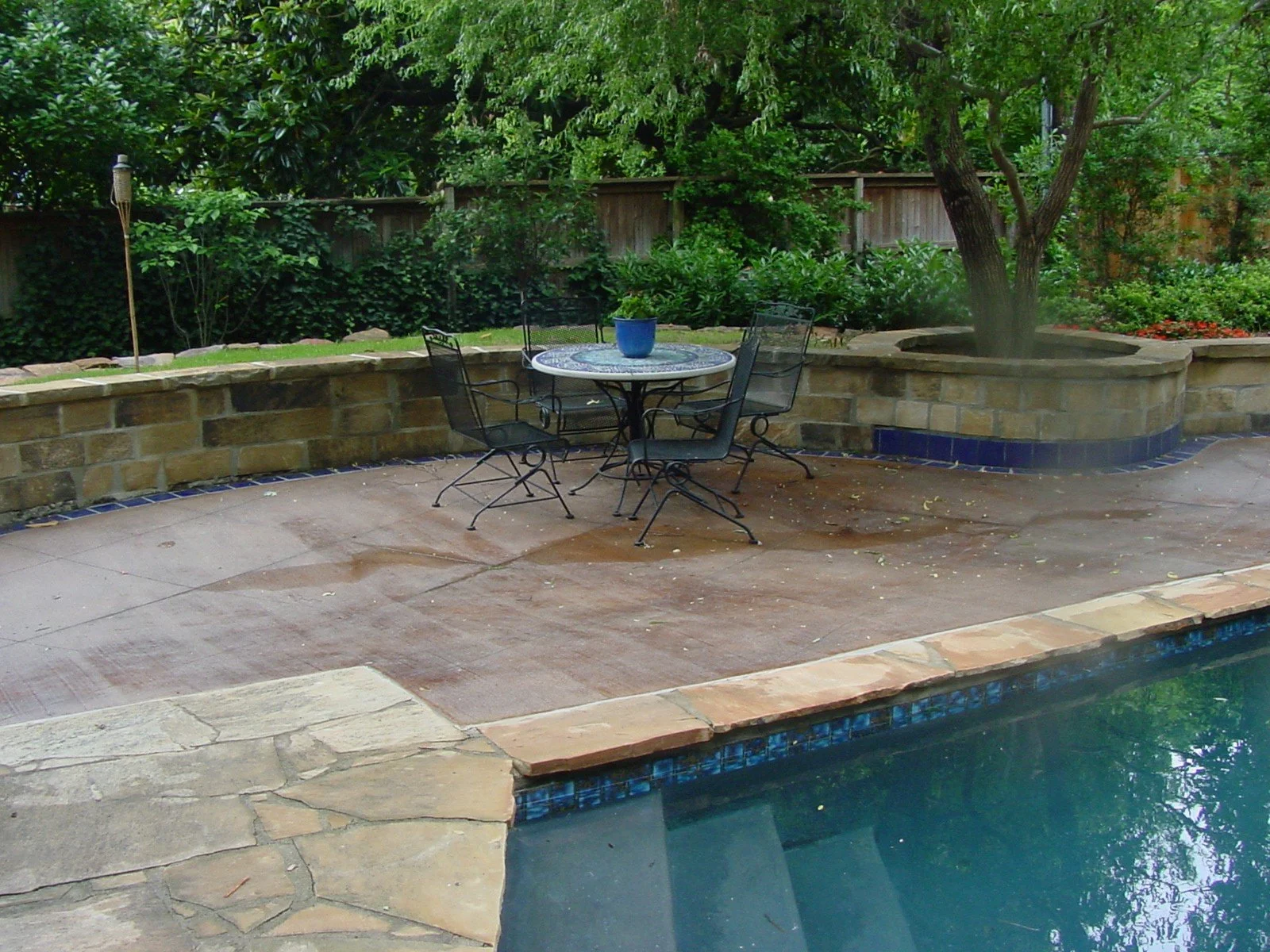 A backyard patio area with a swimming pool in the foreground, a round table with four chairs and a blue planter in the center, a landscaped garden with trees and bushes, and a wooden fence in the background.