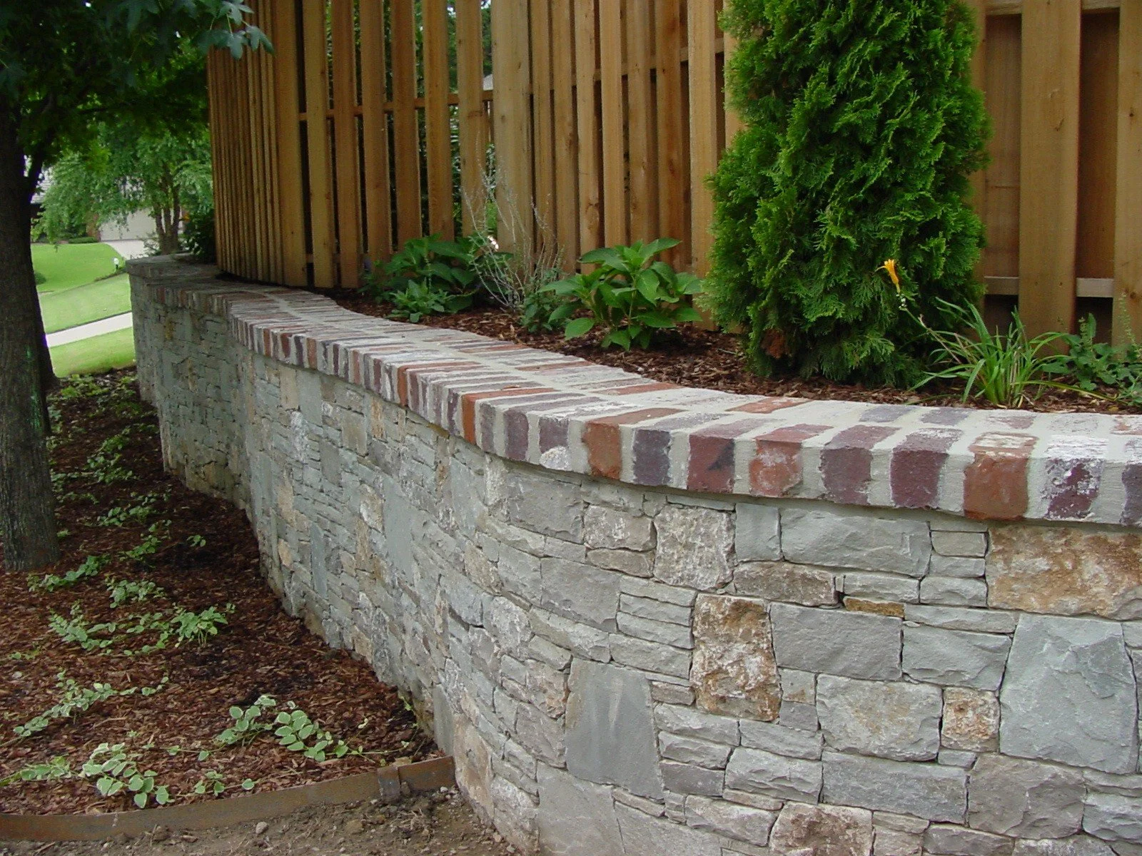 A curved garden with a stone retaining wall topped with a brick border, wooden fence, green plants, and trees in the background.