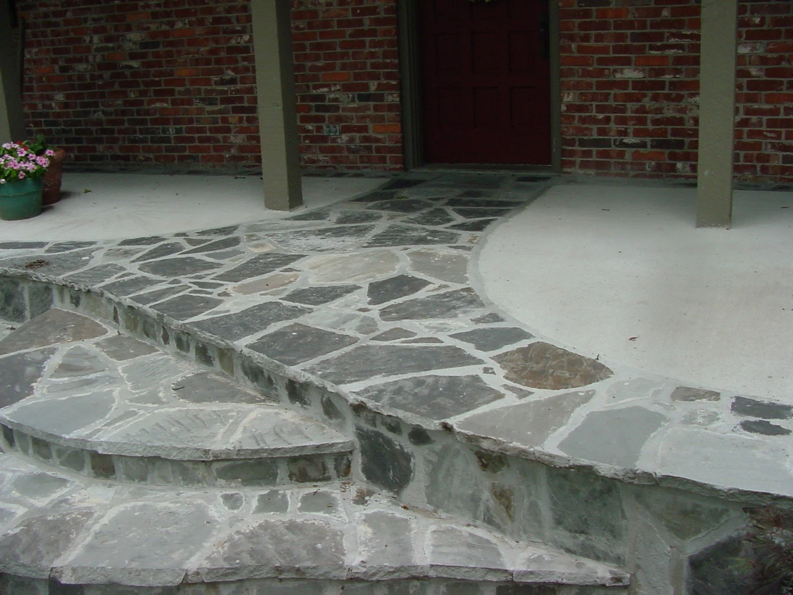 Porch area with stone pathway and steps leading to a door, brick wall background, plants in pots on the left.