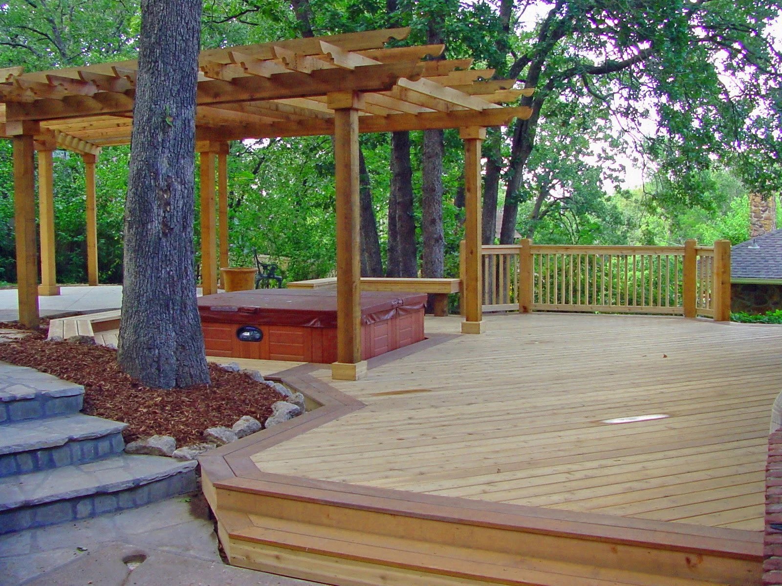 A wooden deck with a hot tub, trees, and a pergola under construction in a backyard.