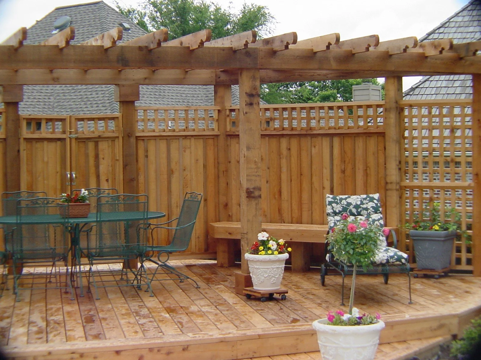 A wooden outdoor deck with a metal table and chairs, potted flowers, and a cushioned chair against a wooden privacy fence.