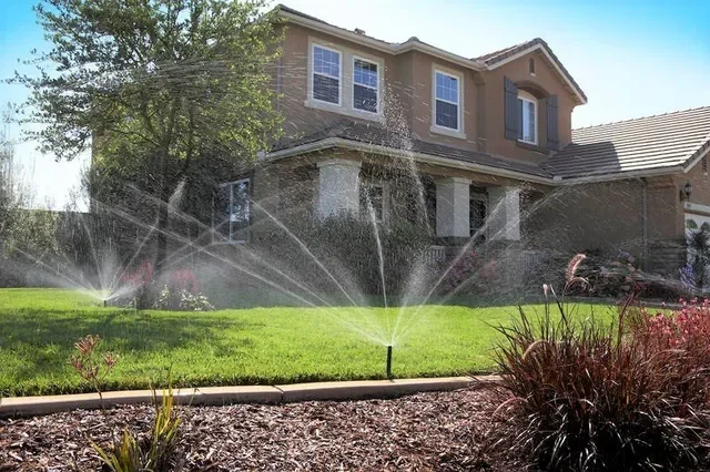 A two-story house with a lawn and garden, with a sprinkler watering the grass in the front yard.