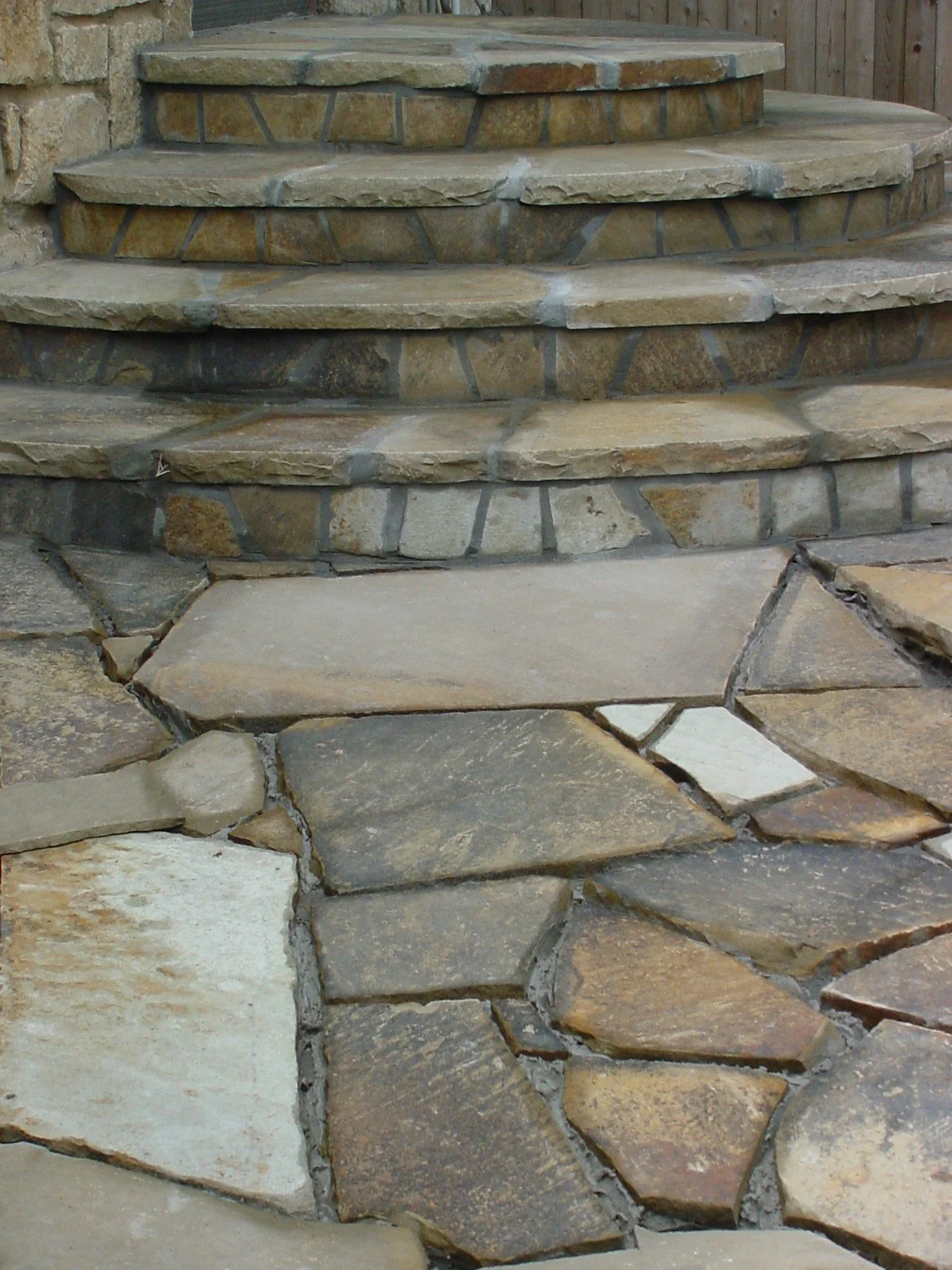 Close-up of a stone staircase and stone pathway with irregularly shaped stones, set against a brick wall and wooden fence.