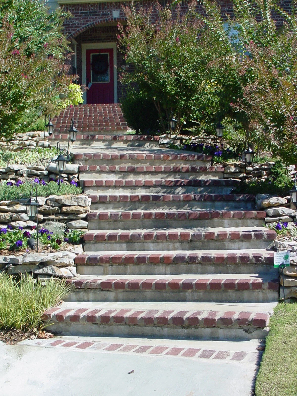 Brick stairs leading up to a house surrounded by plants and flowers, with small lamps along the sides.