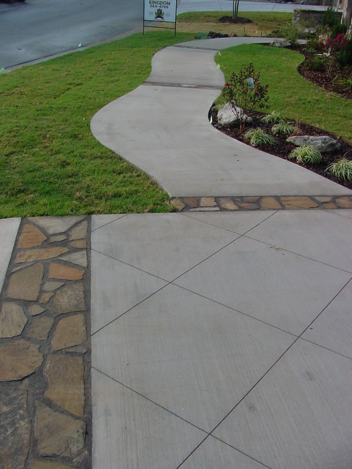 A concrete sidewalk with a curved shape, bordered by grass and landscaped flower beds with plants and large stones, in front of a street with a sign visible in the background.