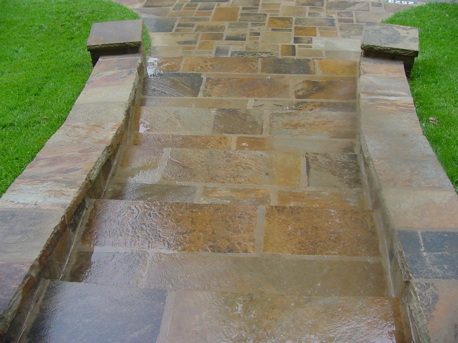 Close-up of a wet, multi-colored stone pathway with raised stone edges, surrounded by green grass.