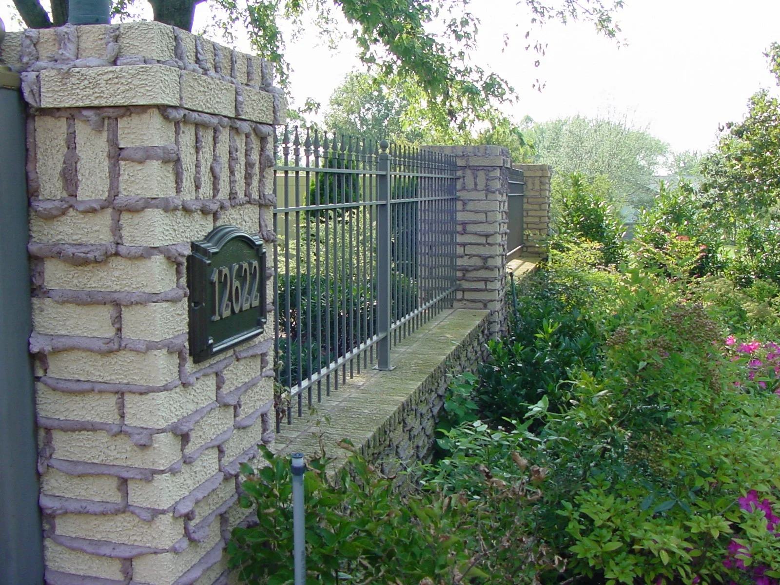 A stone and metal fence with a house number plaque, surrounded by lush greenery and flowering plants.