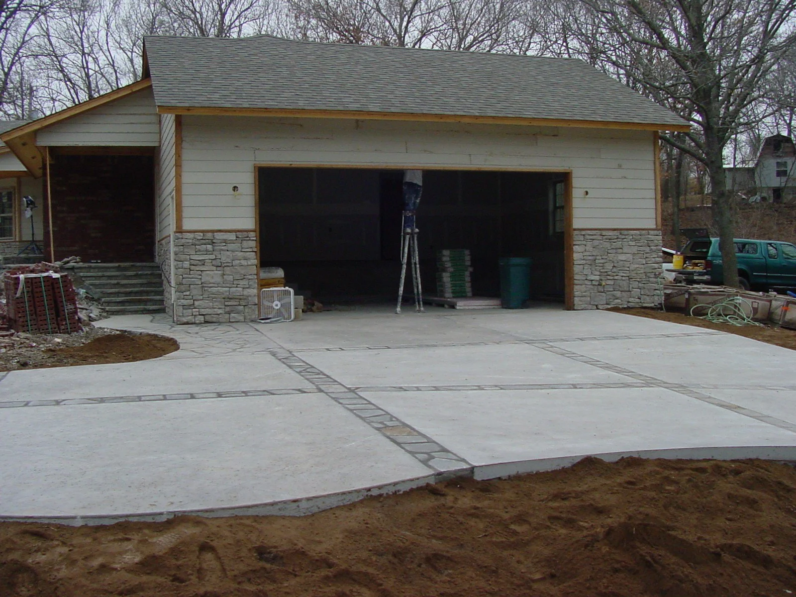 A house under construction with a new concrete driveway and a garage opening, construction materials scattered around, and a tree to the right.
