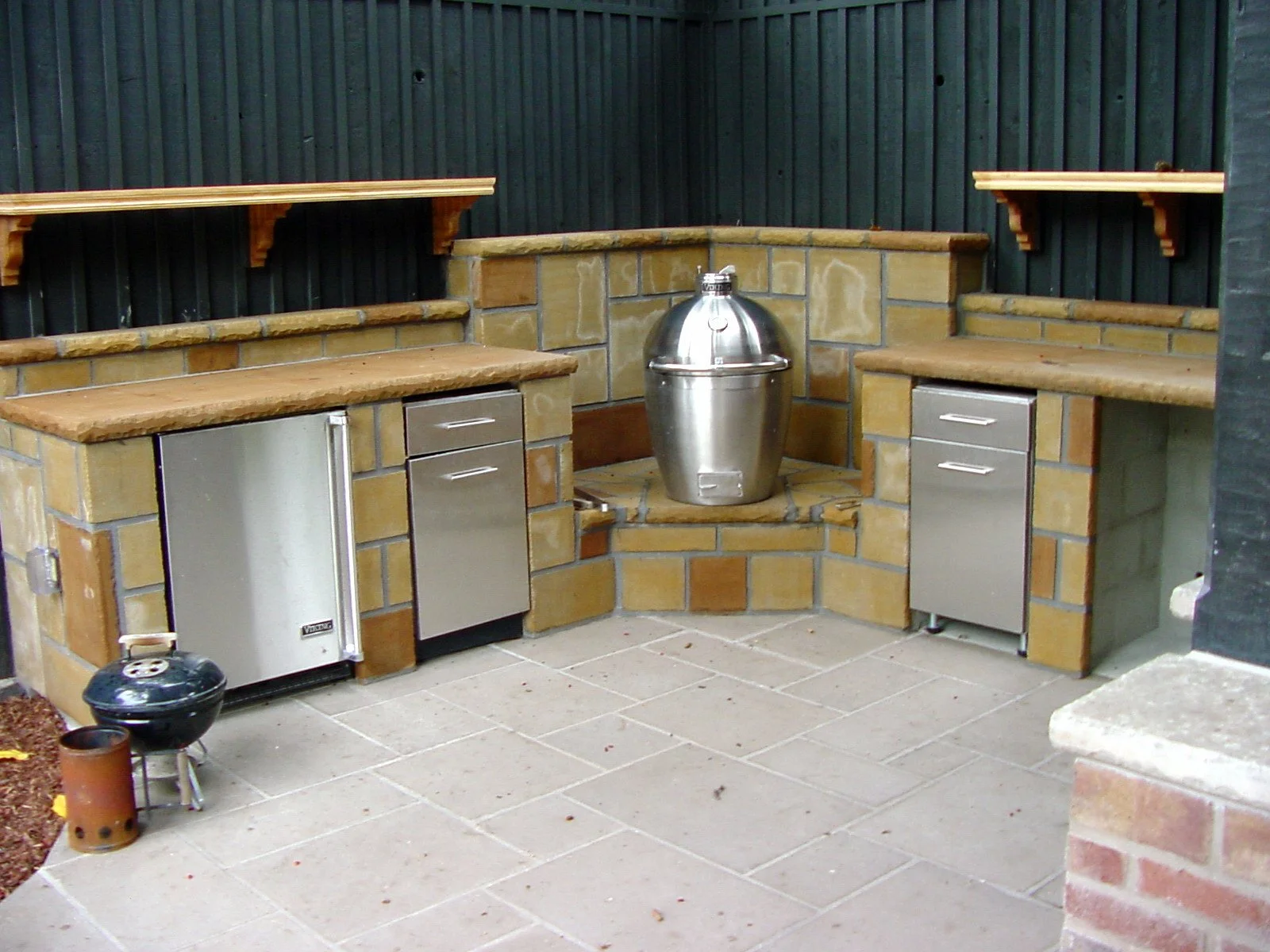 Outdoor bar area with brick and wood countertops, stainless steel mini-fridges, a metal beer keg in the center on a brick stand, and small shelves above the countertops. The area is paved with large light-colored tiles.