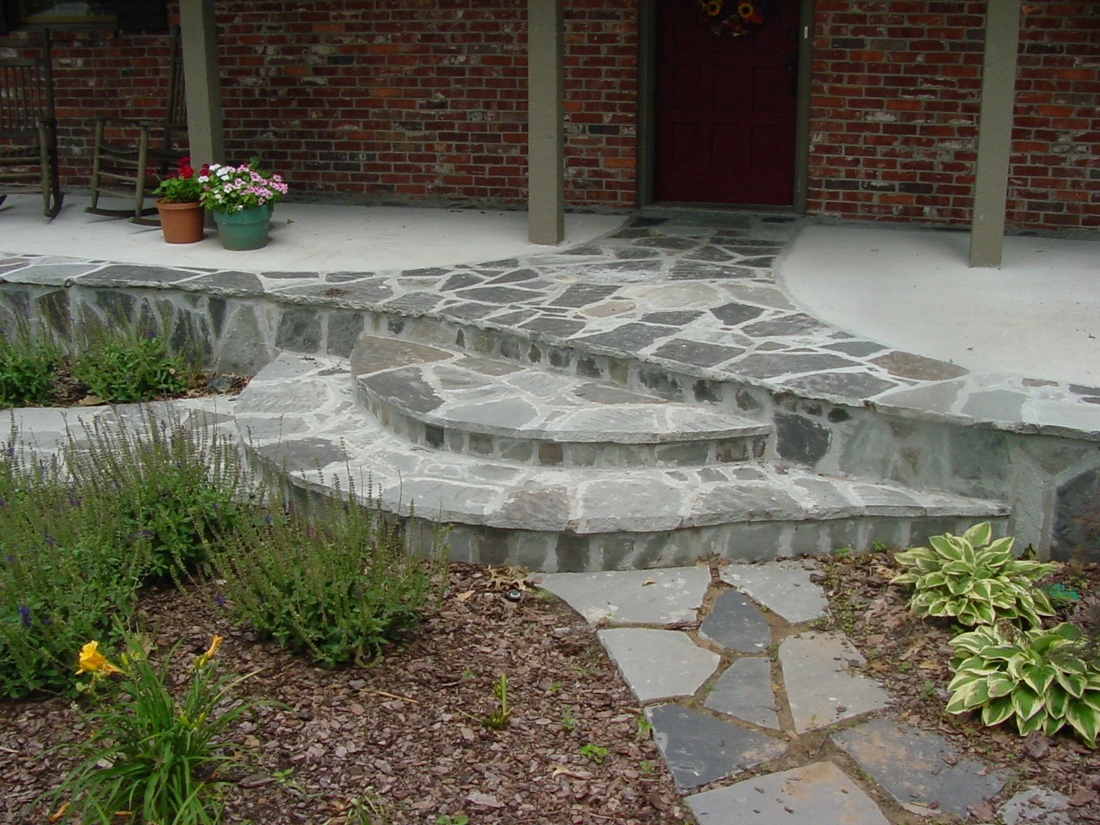 A brick house porch with stone steps leading to the front door, adorned with potted flowers and surrounded by garden plants.