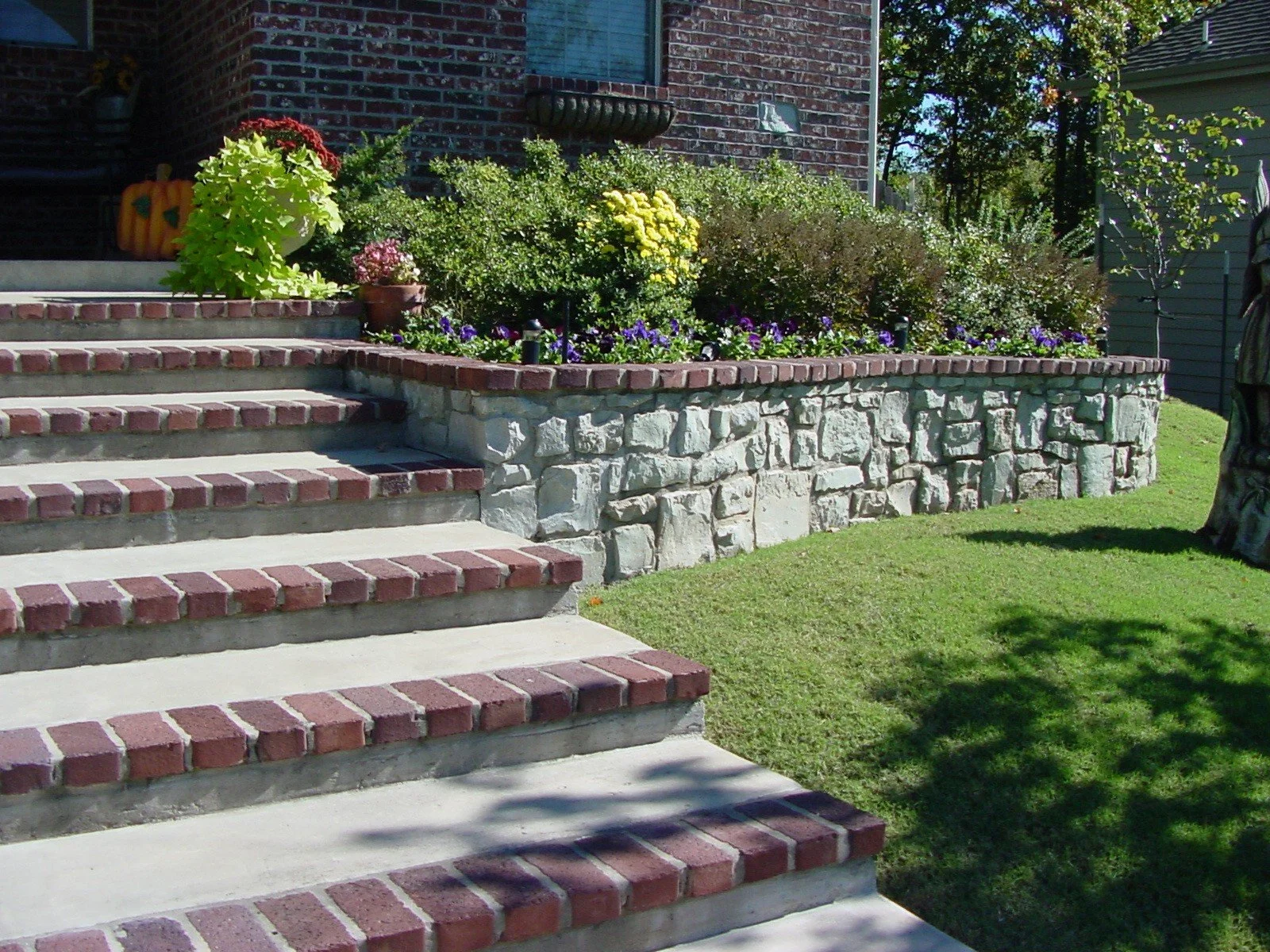 Brick stairs leading up to a house with a flower bed and stone retaining wall, green lawn, and shadows on the grass.