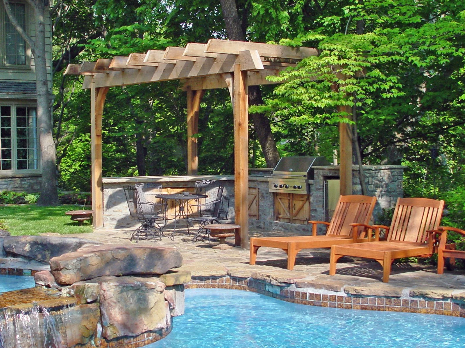 Backyard patio with a swimming pool, wooden lounge chairs, and a covered outdoor kitchen area with a grill, surrounded by lush green trees.