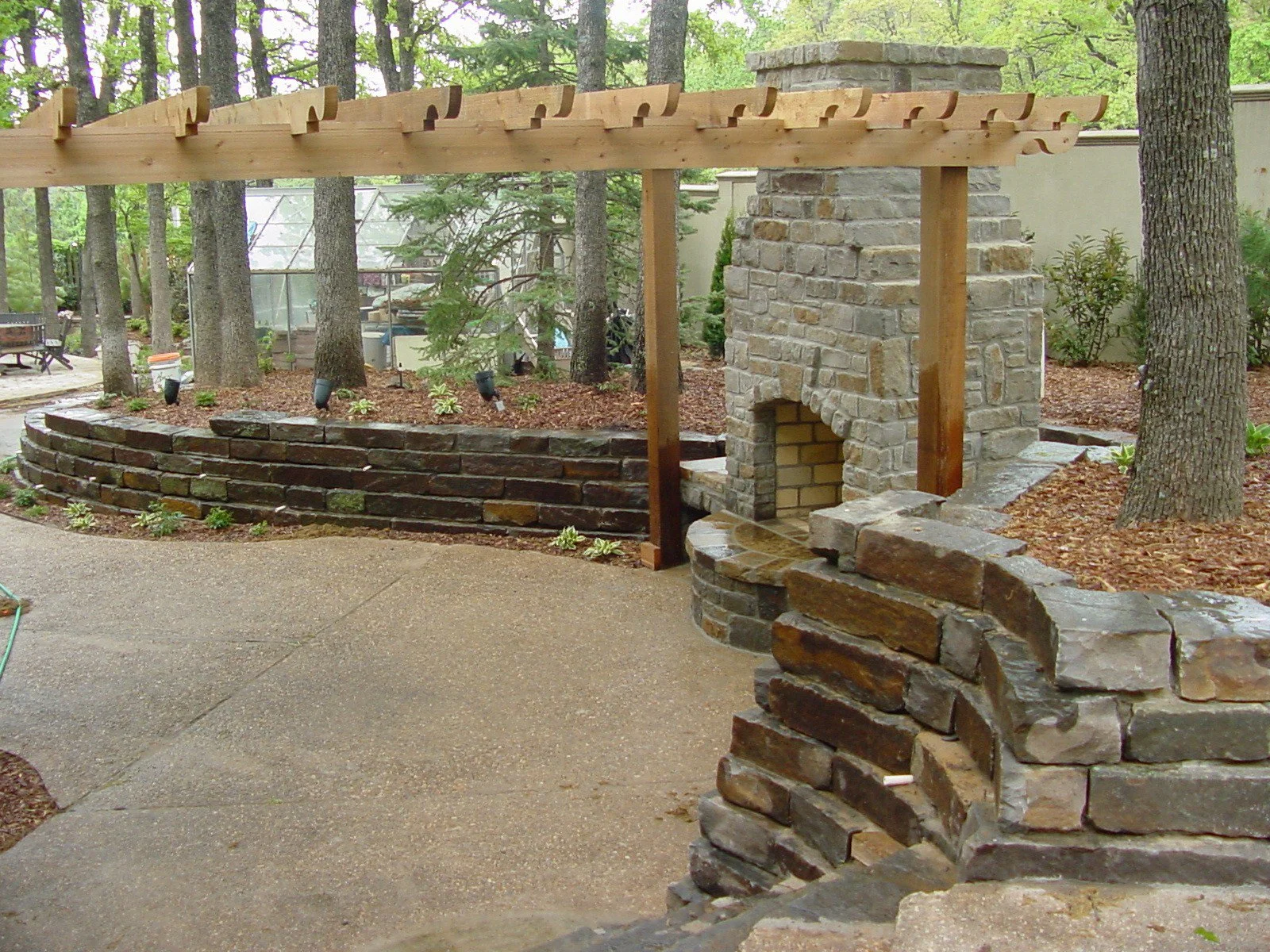 Backyard with a stone fireplace, brick and stone retaining walls, and a wooden pergola surrounded by trees and plants.