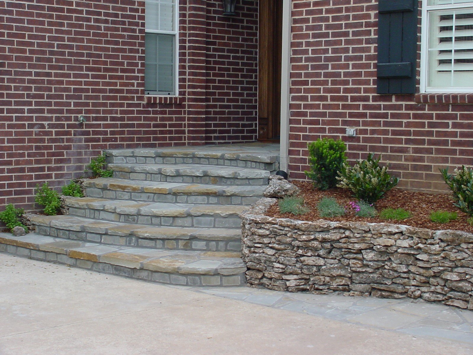 Red brick house entrance with stone steps leading to a wooden door, landscaped with shrubbery and stone retaining wall.