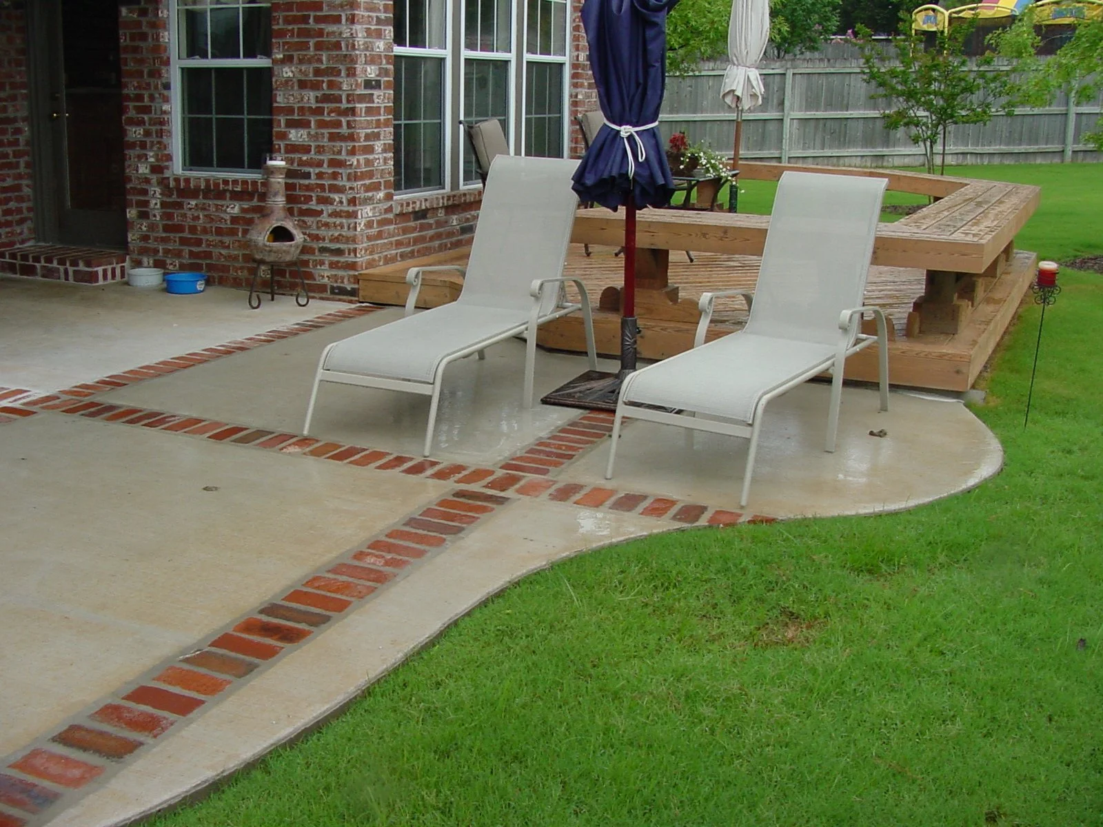 Backyard patio with two white lounge chairs, a closed blue patio umbrella, a small wooden deck, a brick chimney, a brick and concrete pathway, and a fenced green lawn with small trees.