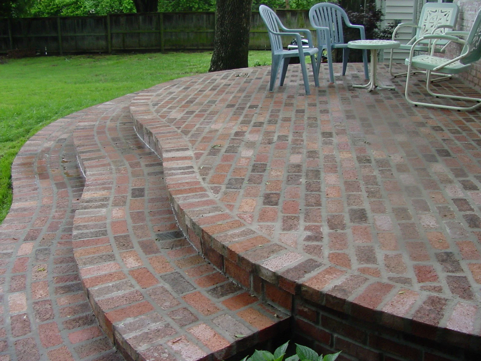 Photo of a curved brick patio with outdoor chairs and a small table, surrounded by green grass and a wooden fence in the background.