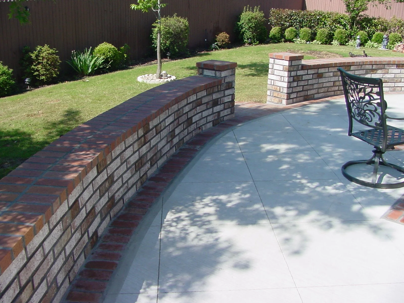 Backyard patio with a curved brick wall, concrete flooring, and metal chair; green lawn, plants, and trees in the background with a wooden fence.