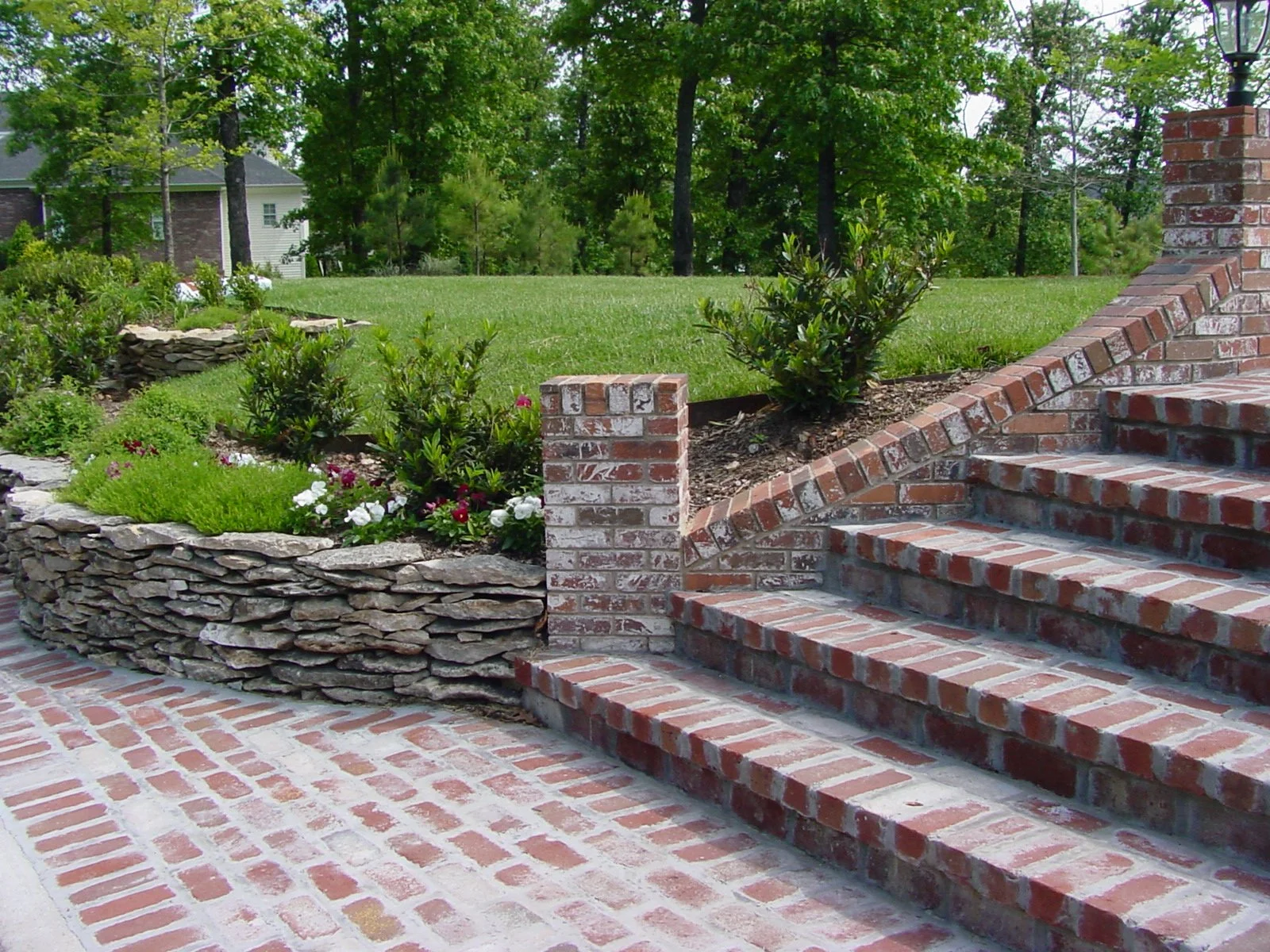 Brick steps leading up to a landscaped yard with a stone retaining wall and flowering plants, surrounded by green trees.