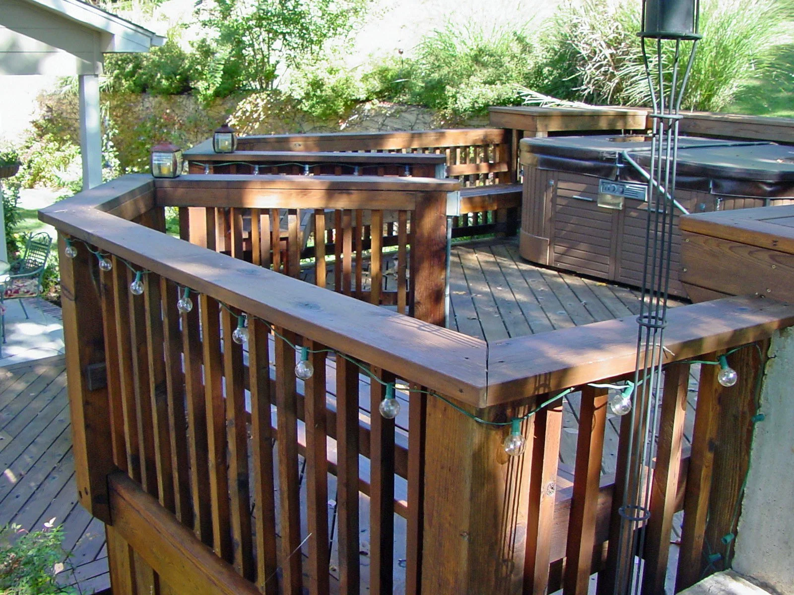 Wooden deck with railing, string lights, patio furniture, and a hot tub surrounded by greenery.