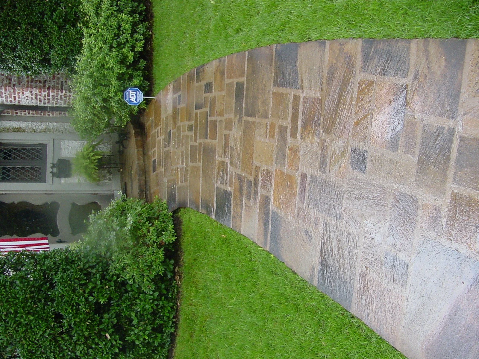 A wet stone pathway leading up to a house with a front porch. There is a small American flag hanging on the porch and bushes along the walkway. An 'ADT' security sign is also visible near the bushes.