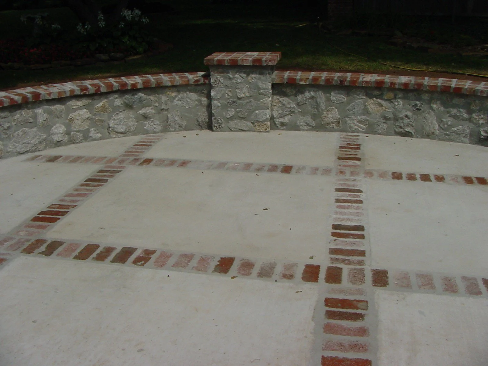 Concrete patio with brick inlay patterns, stone wall with brick cap, and a grassy yard in the background at night.