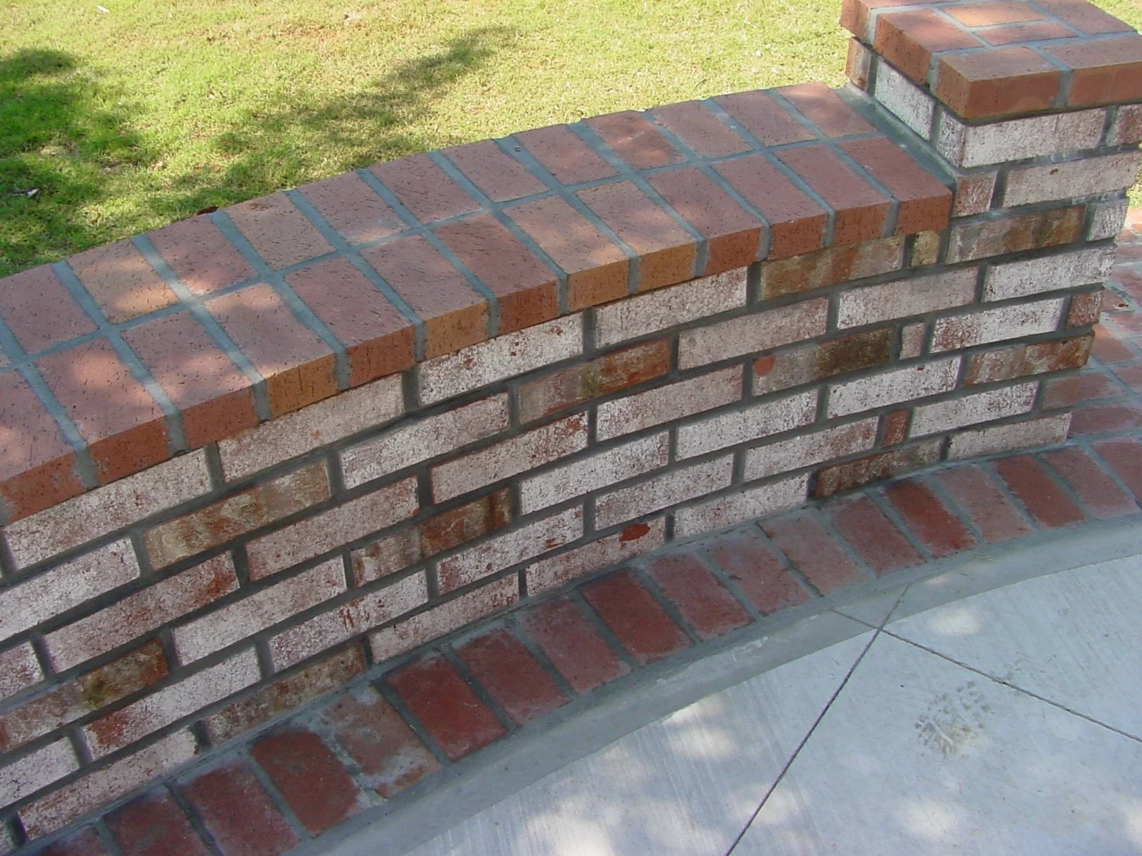 Close-up of a brick wall with a curved concrete border, background shows grass and a sidewalk.