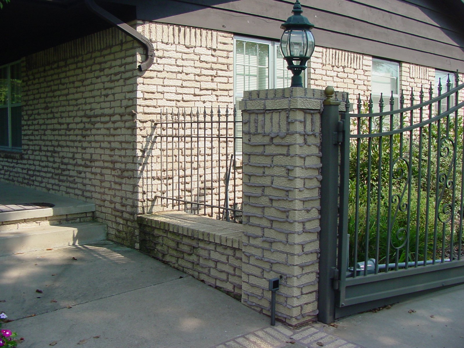 A brick wall and a black wrought iron gate with stone pillars, a lamp post on top of one pillar, and greenery in the background.
