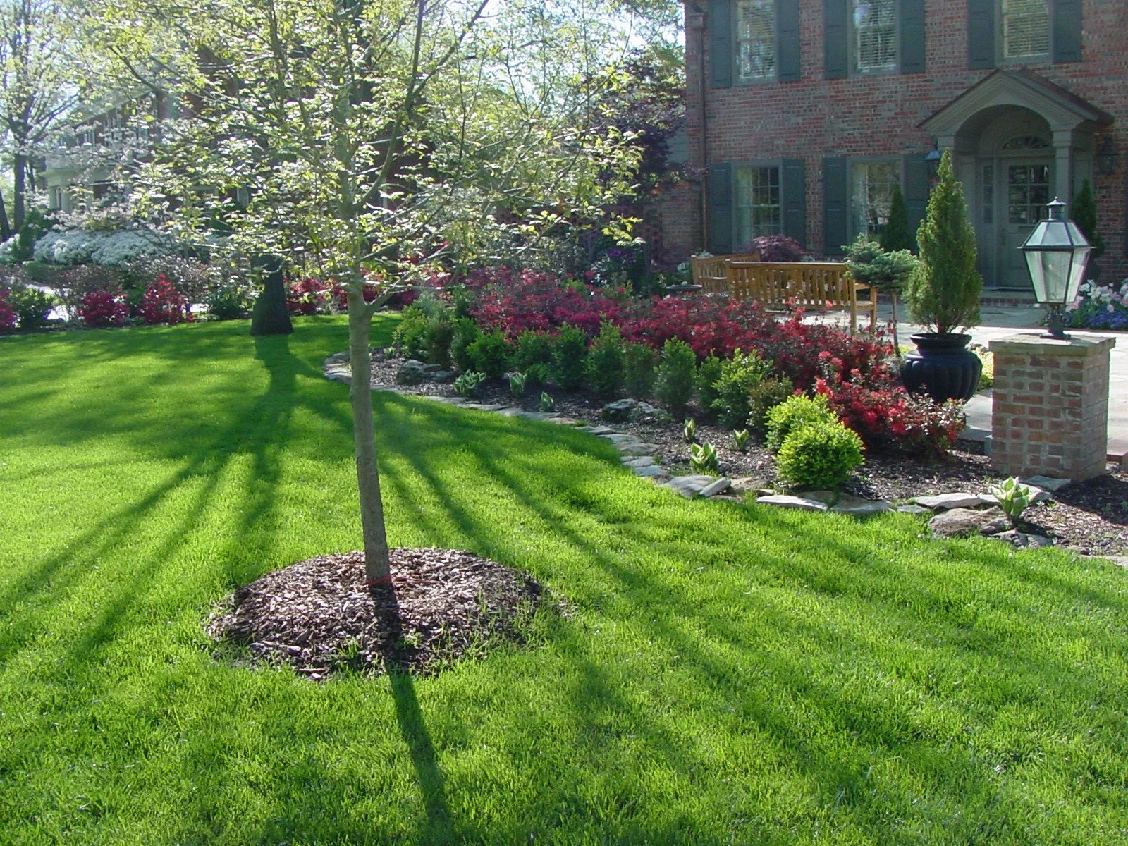 A well-maintained front yard garden with green grass, a small tree, flowering bushes, decorative plants, and a brick house in the background. There is also a stone border and a lamp post on the porch.