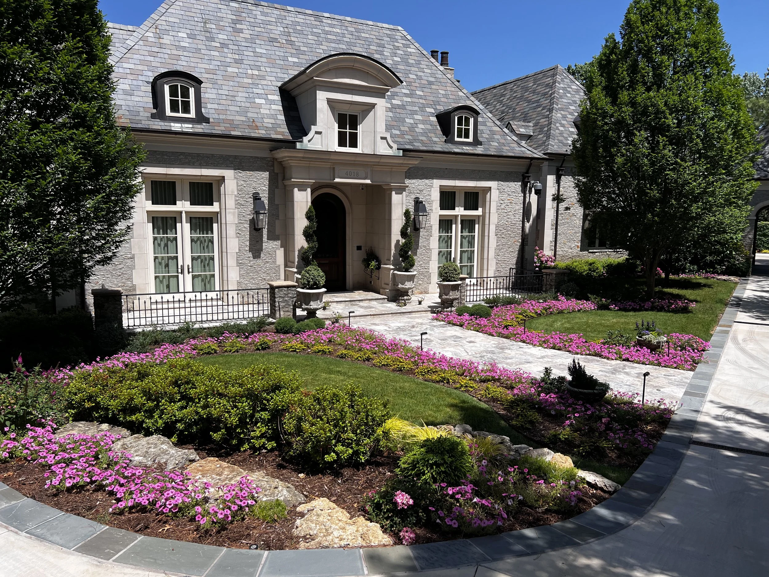 A house with a slate roof and stone exterior, surrounded by a landscaped garden with flowering plants and small trees, with a curved pathway leading to the front entrance on a sunny day.