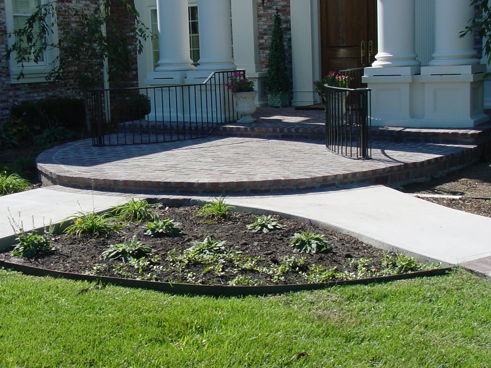 Front porch of a house with a brick steps, white columns, black railing, potted plants, and a garden in the foreground.