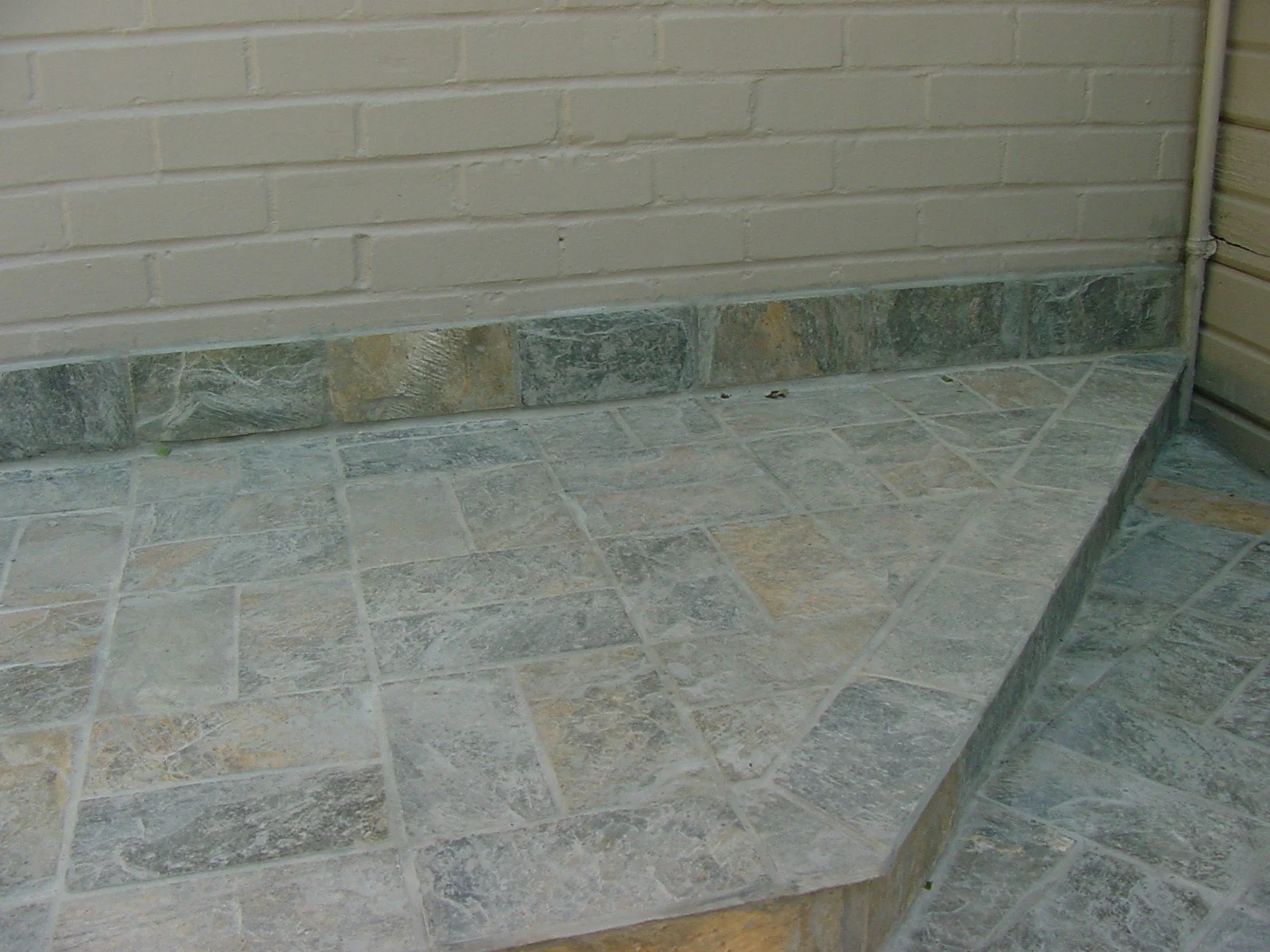 The image shows an outdoor corner with beige brick wall and stone tiles on the ground and a step made of similar stone tiles.