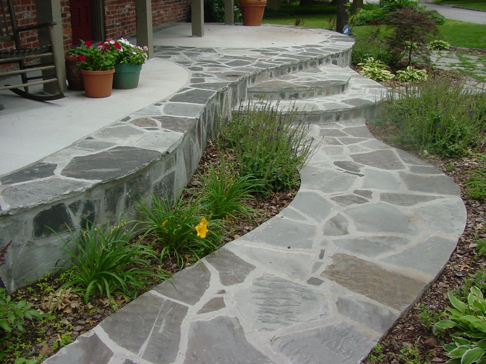 A stone pathway with curved and irregularly shaped stones leading to a porch with potted plants, surrounded by a garden with various green plants and flowers.