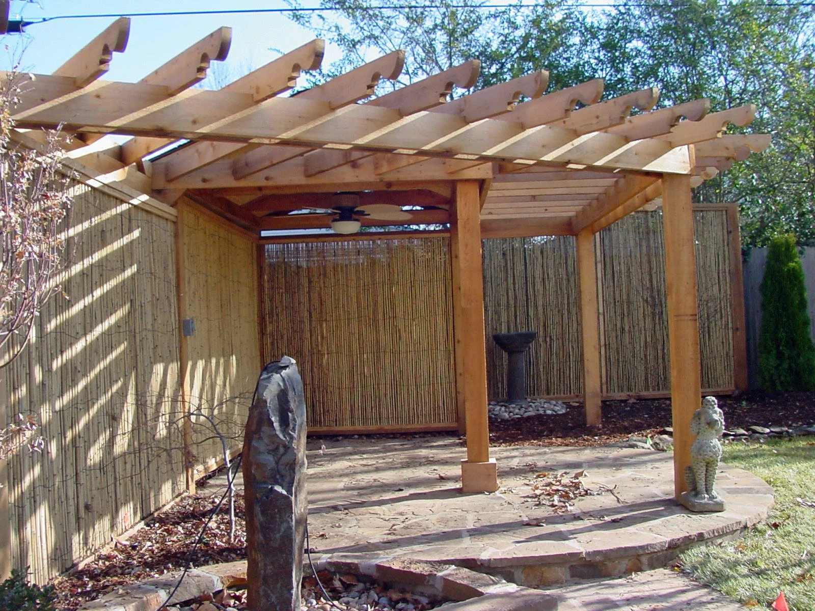 Backyard with wooden pergola, bamboo privacy fence, garden statues, and a birdbath. The yard has a paved patio area and some trees in the background.