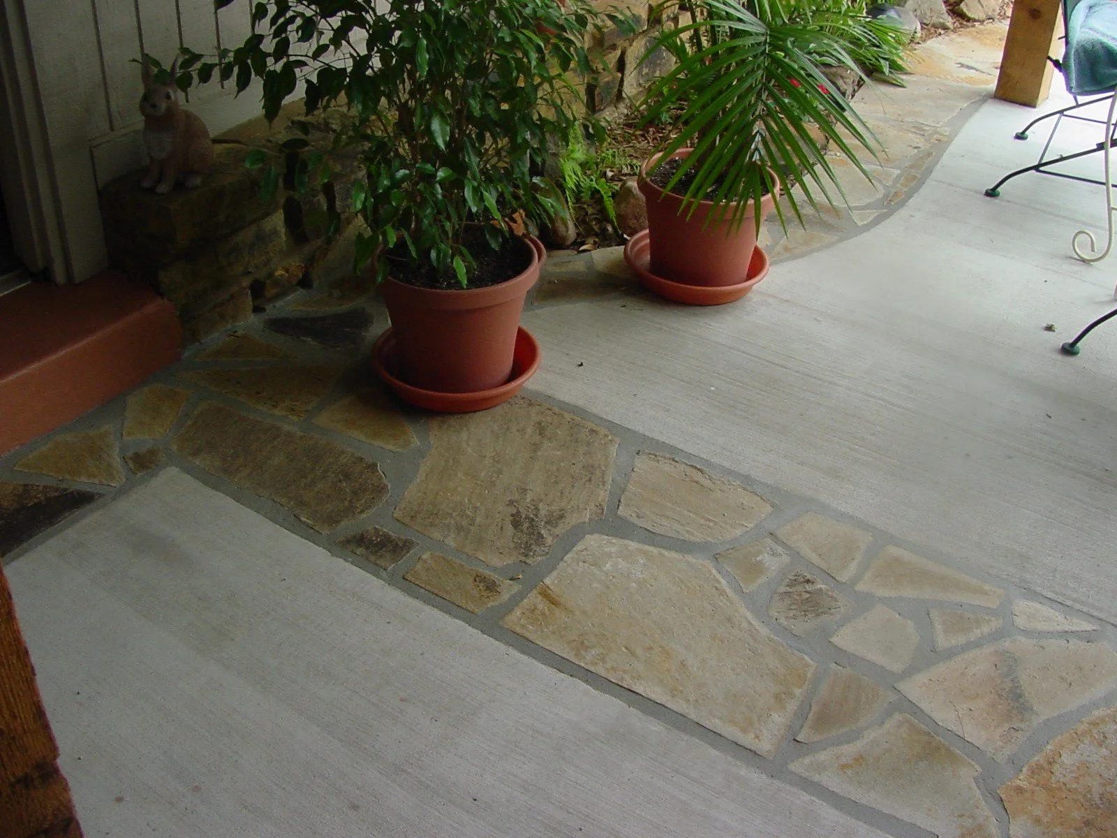 A porch with potted plants on a concrete and stone floor, including two large terracotta pots with green leafy plants, and a small rabbit figurine on a stone step.