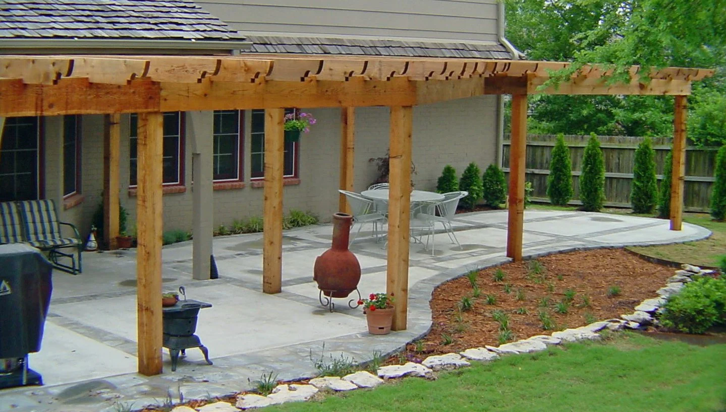 Backyard patio with a covered wooden pergola, outdoor furniture including a table and chairs, a chiminea, potted plants, landscaped flower beds, and a wooden fence with shrubs.