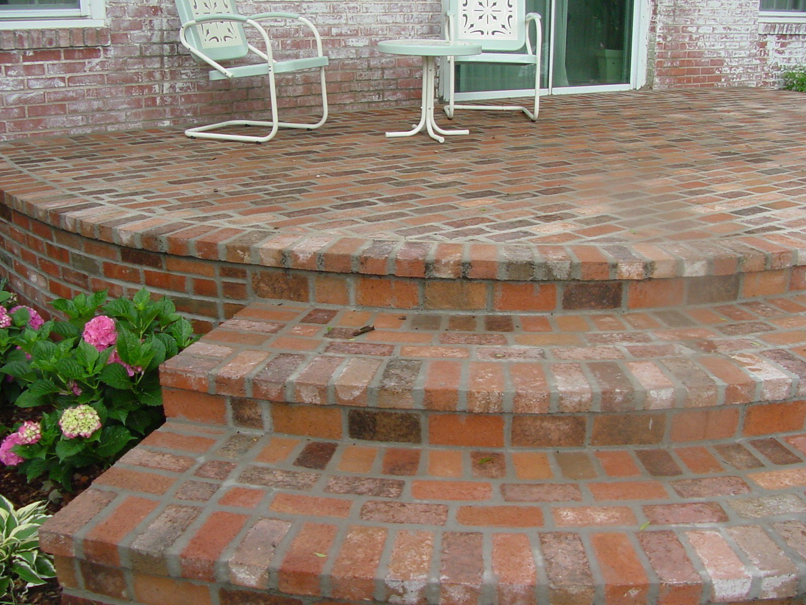 A brick patio with three wide steps, a small round table, two chairs, a sliding glass door, and a flower bed with pink hydrangeas.