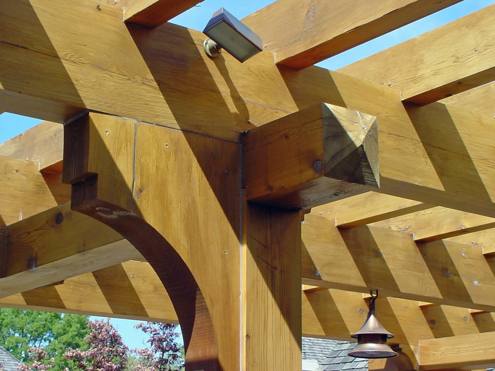 Close-up of a wooden pergola structure with beams and a small outdoor hanging light fixture, trees and rooftops visible in the background.