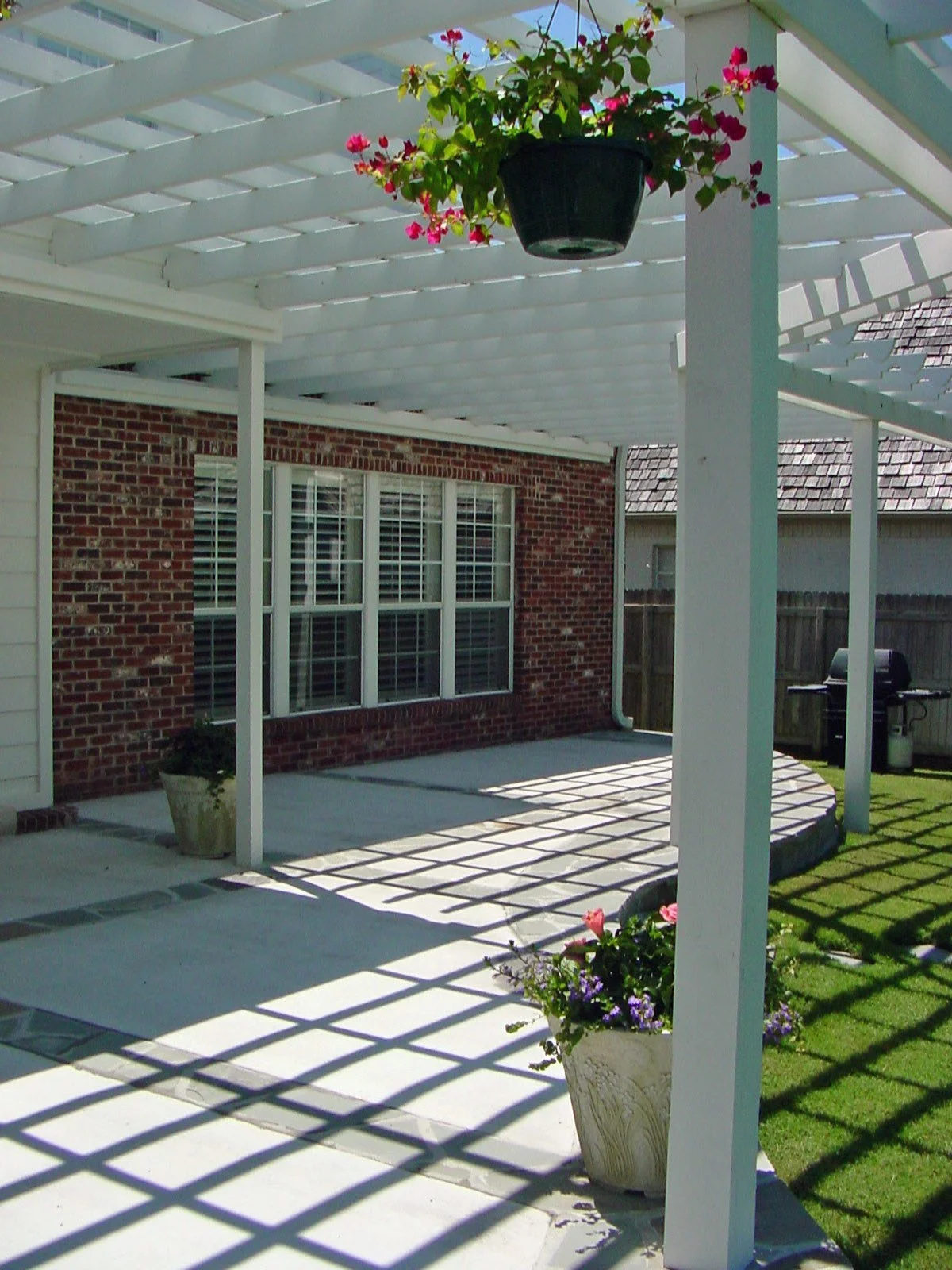 Backyard patio with a white pergola, potted flowers, and a brick house with large windows.