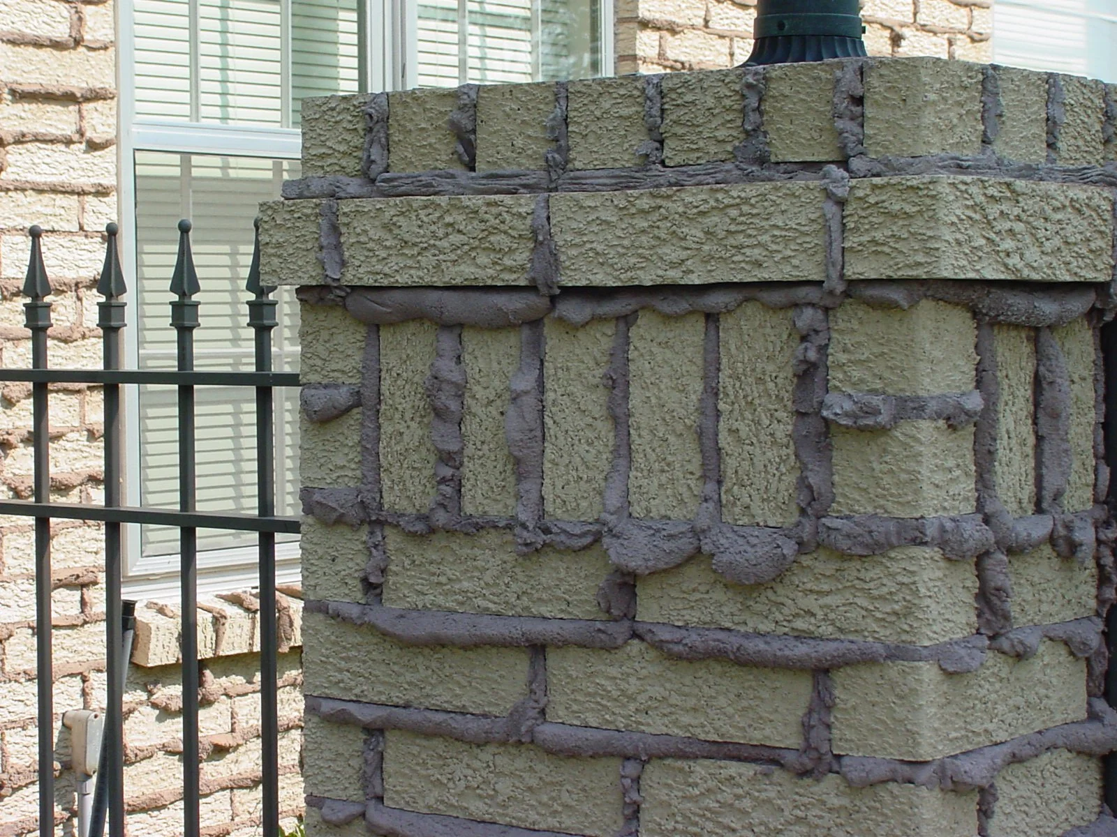 Close-up of a brick column with mortar applied for joint filling, next to a brick wall with a window and decorative metal fence in the background.
