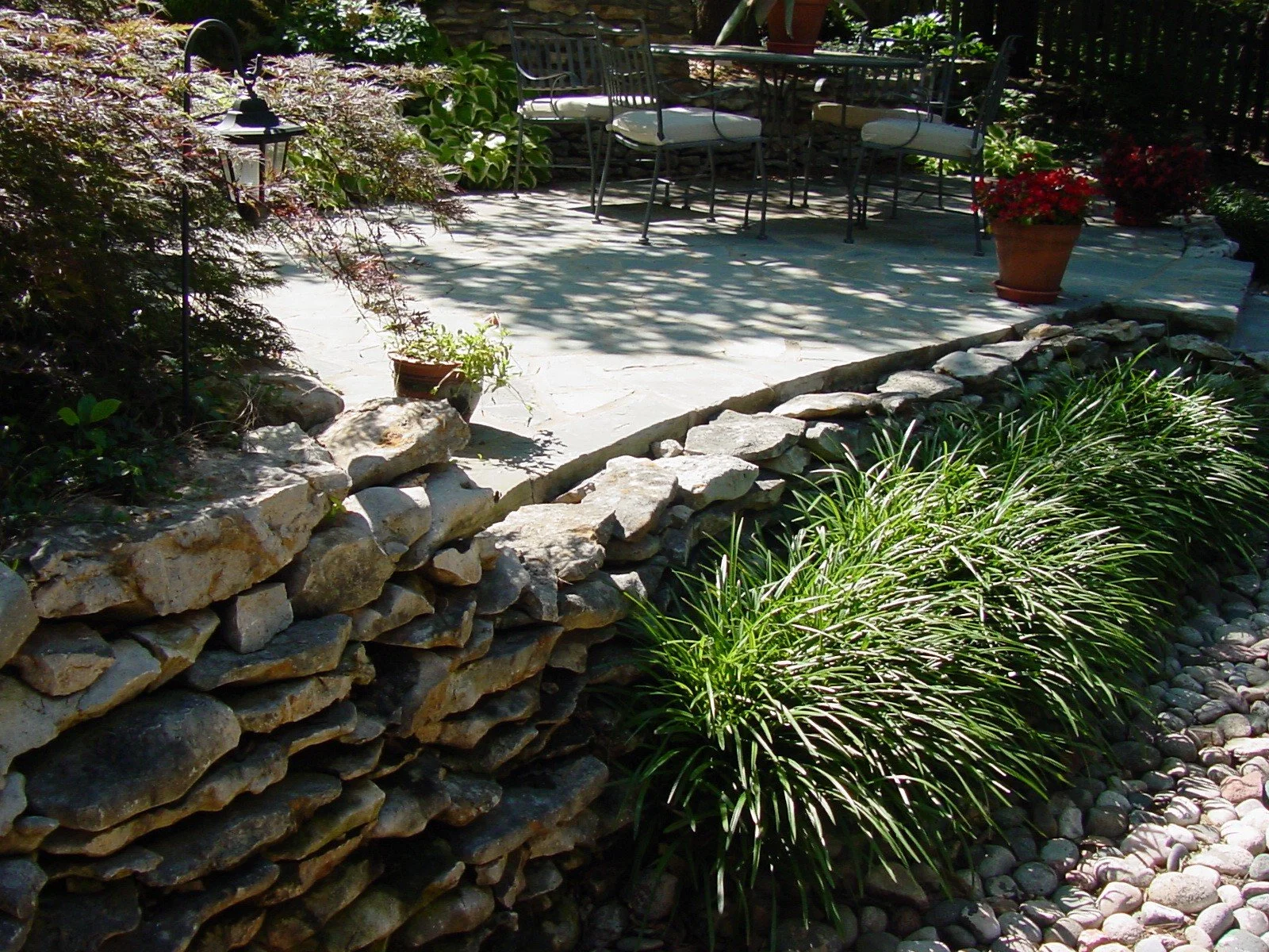 A garden patio with a rock border, potted plants, and outdoor furniture including chairs and a table, surrounded by greenery and shaded by trees.