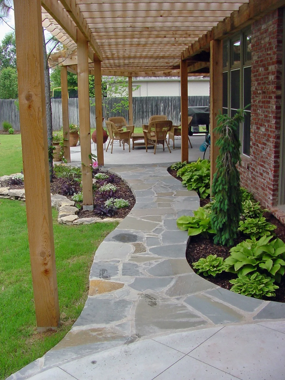 A backyard patio with a stone pathway, garden beds, and an outdoor dining area with chairs and a table under a wooden pergola.